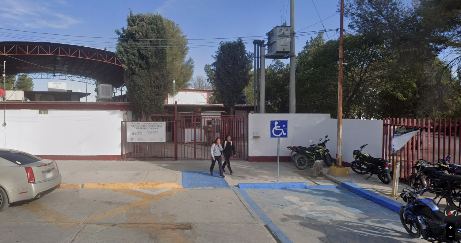 Fotografía en la que se muestra la entrada del plantel, que está conformada por muros blancos y una reja color rojo por la que se puede ingresar a las instalaciones. Frente a la reja hay una rampa de acceso. 